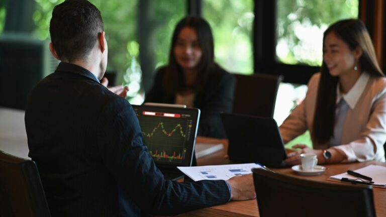 One man in a suit looking at a laptop screen with data projections, sitting across from him are two professionally dressed women at a conference table with papers in front of them.