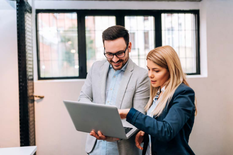 Two business professionals standing and collaborating while looking at a laptop screen, with a bright office window in the background.