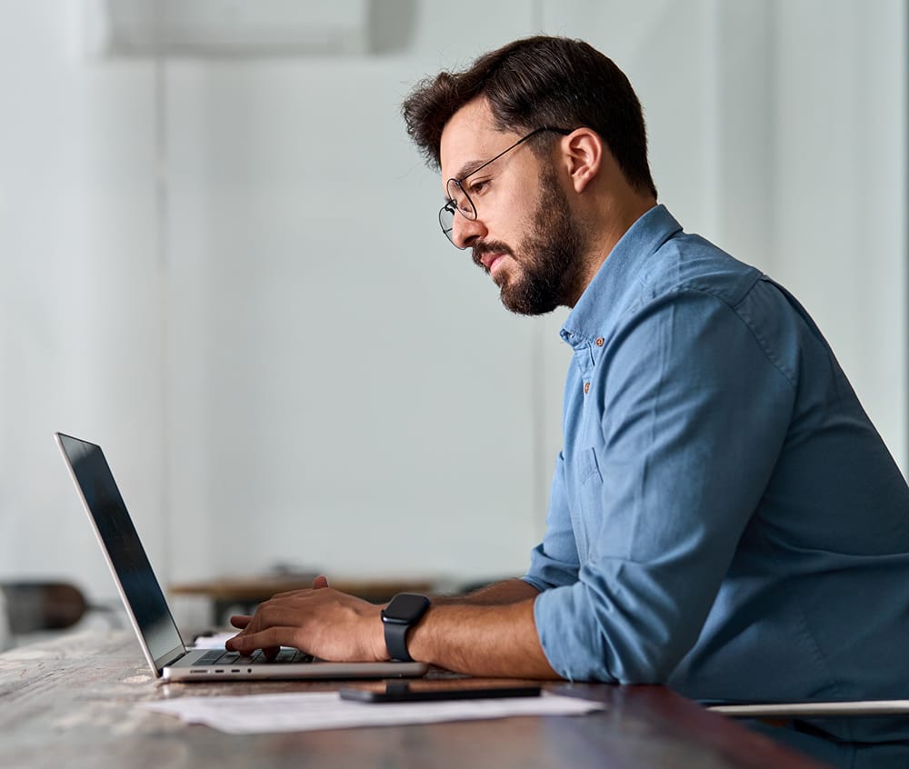 Man in glasses working on a laptop at a desk, focused and engaged in a modern office setting.