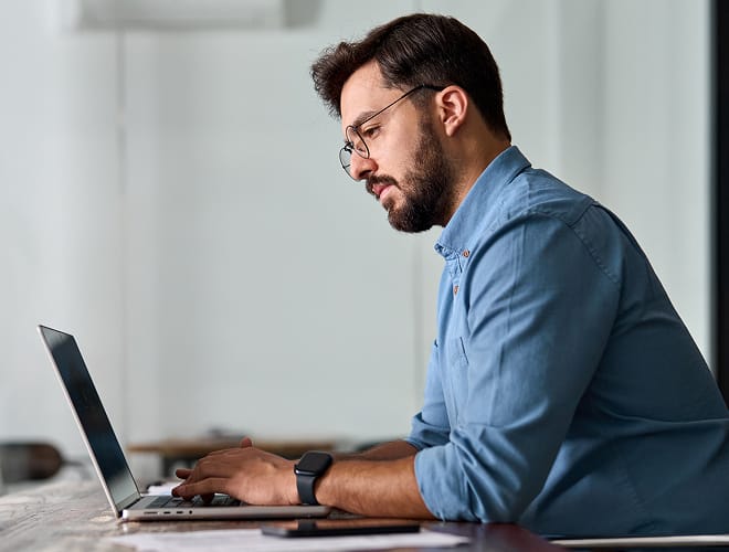 Man in glasses working on a laptop at a desk, focused and engaged in a modern office setting.