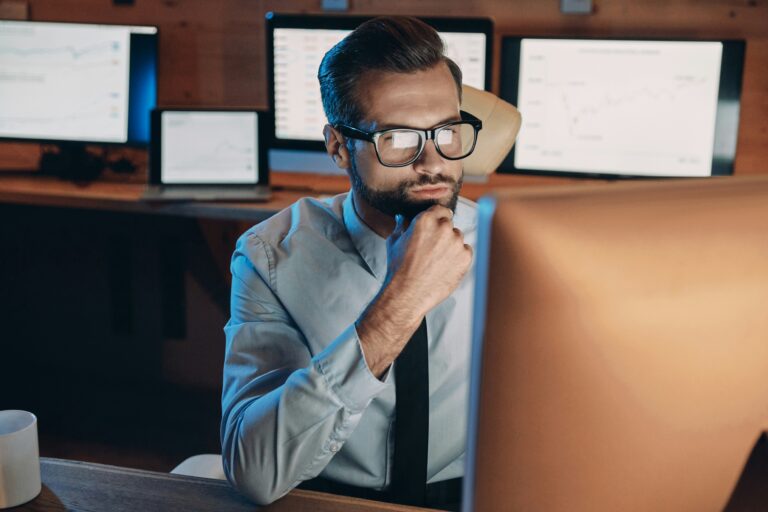 Business professional wearing glasses and a dress shirt, sitting at a desk and concentrating on a computer monitor, with multiple data and graph screens in the background.